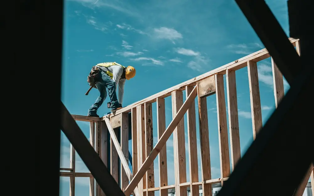 A builder in a hard hat working on the wood frame of a house